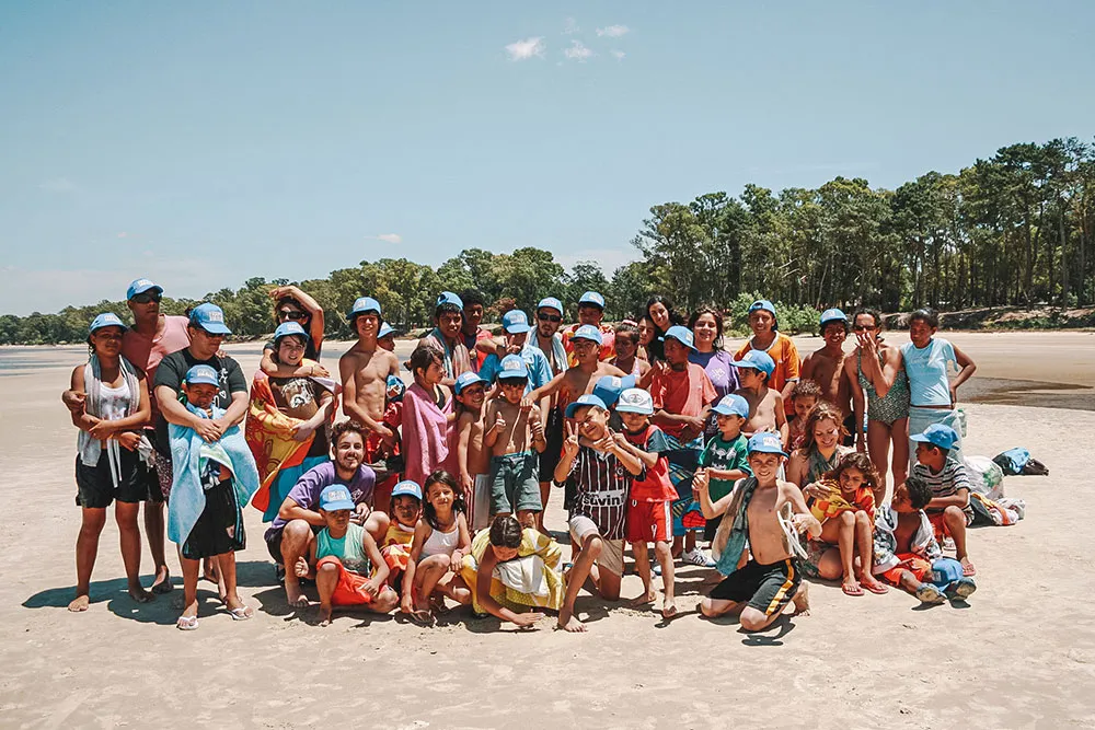 Grupo de voluntários e crianças em uma praia, todos usando bonés azuis e sorrindo juntos.