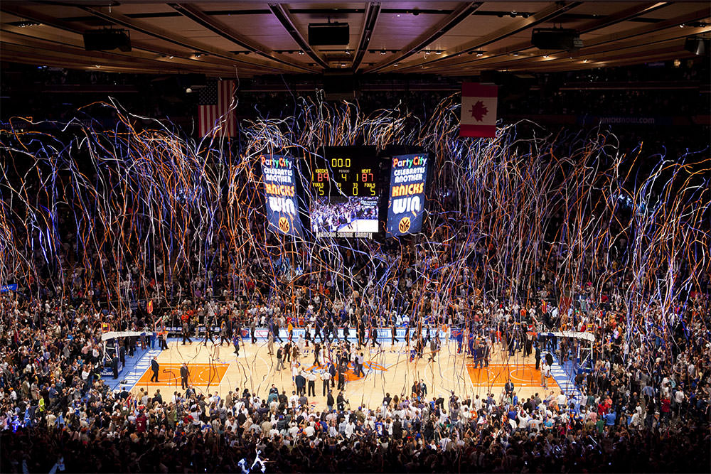 Comemoração de vitória dos Knicks no Madison Square Garden, Nova York, com o público celebrando e serpentinas coloridas no ar.