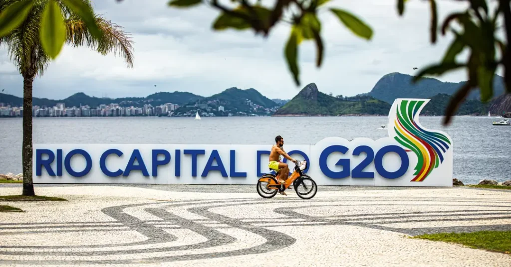 Placa 'Rio Capital do G20' no Parque do Flamengo, com a Baía de Guanabara e o Pão de Açúcar ao fundo.