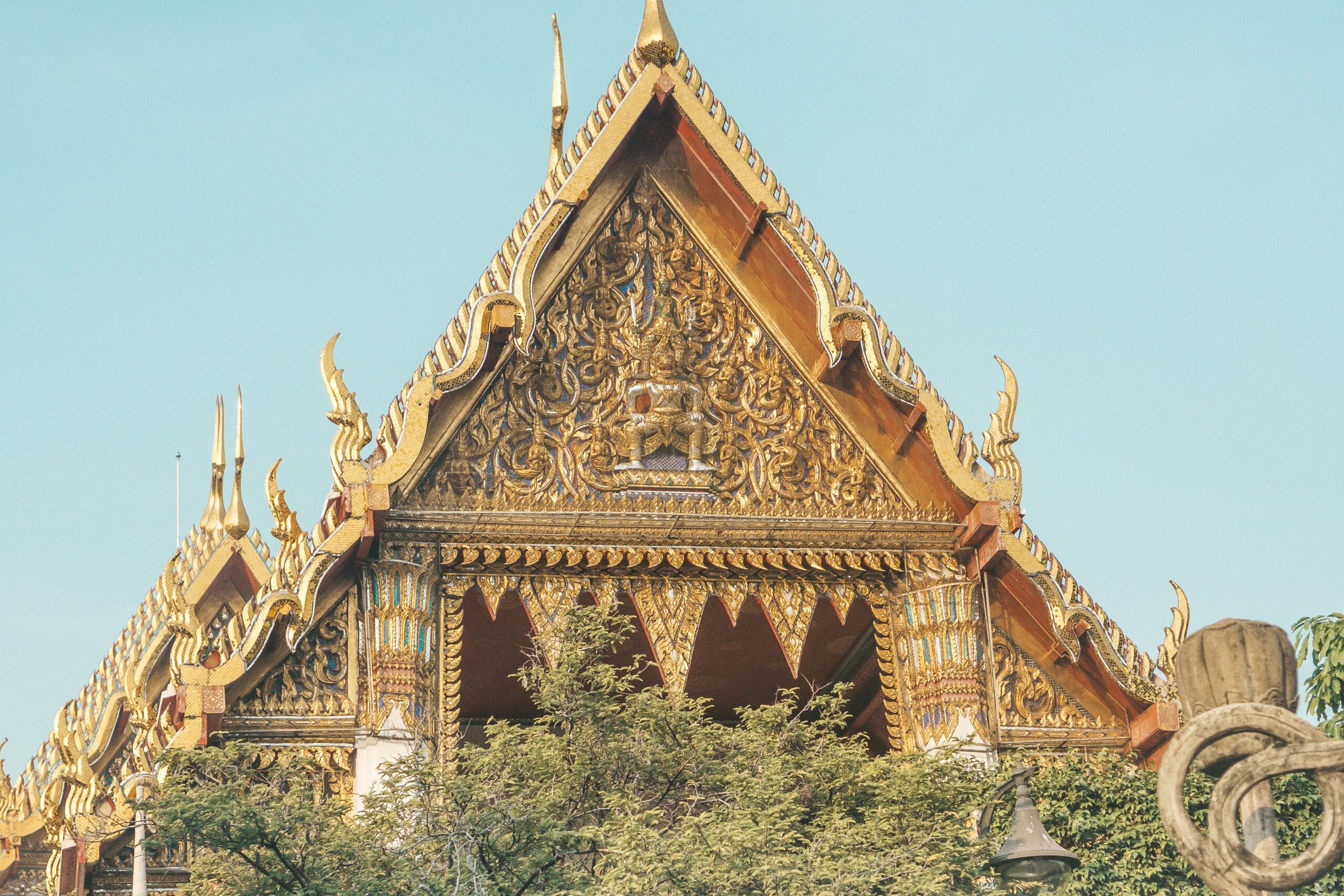 Detalhe arquitetônico de um templo tradicional em Bangkok, Tailândia, com ornamentos dourados e céu azul ao fundo.