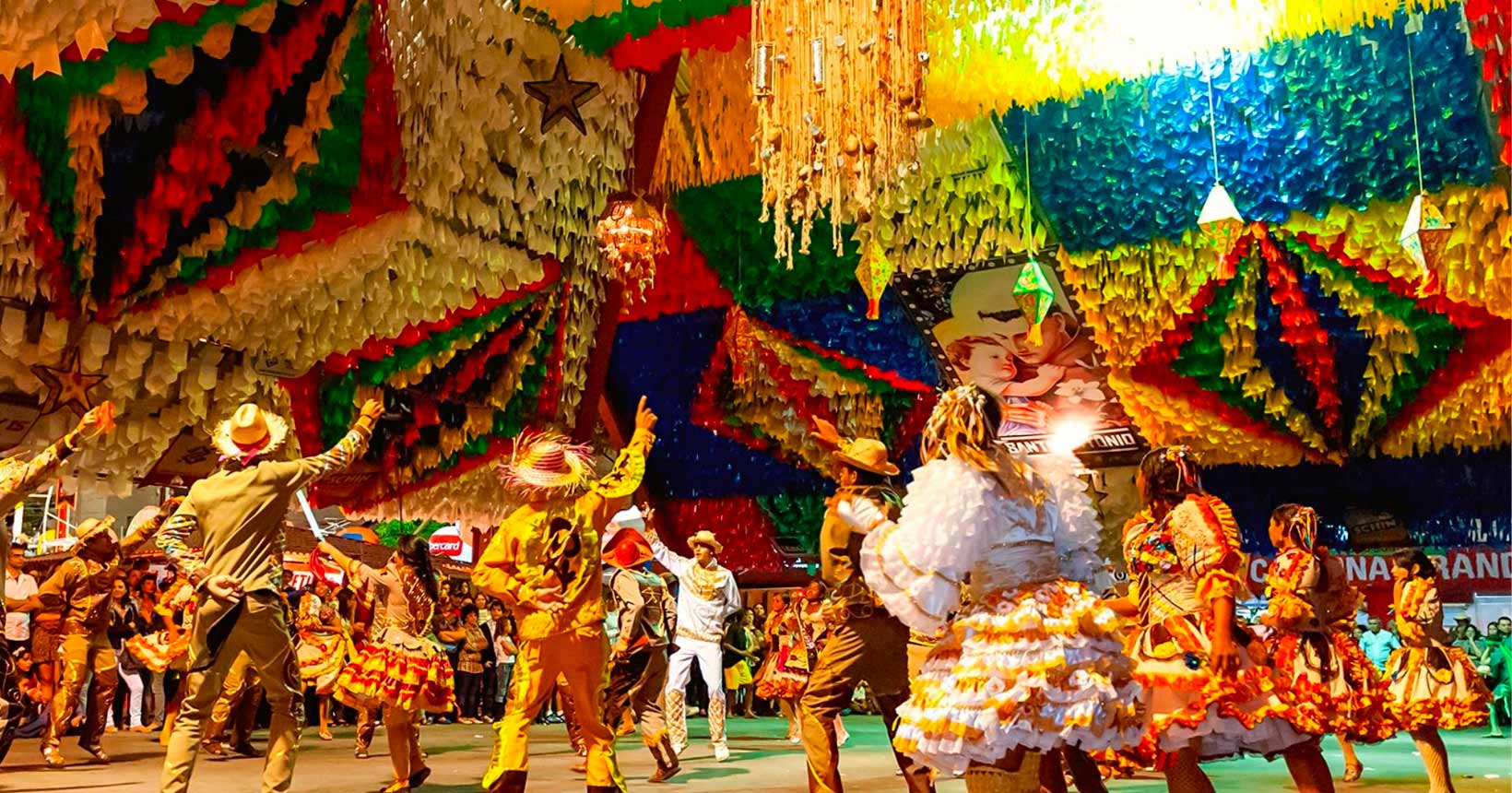 Grupo de dançarinos se apresenta em uma festa típica de Campina Grande, com trajes tradicionais de forró, sob uma decoração colorida de bandeirolas.