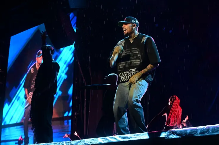 Chris Brown se apresentando no palco durante um show em São Paulo, sob chuva, com dançarinos ao fundo.