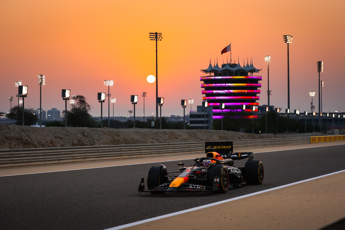 Carro de Fórmula 1 no circuito do GP do Bahrein ao entardecer, com o pôr do sol e a torre icônica ao fundo.