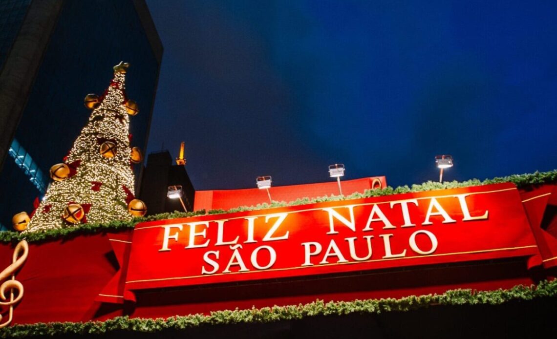 Decoração de Natal em São Paulo com uma grande árvore iluminada e um painel vermelho escrito 'Feliz Natal São Paulo' ao fundo, destacando o clima natalino na cidade.