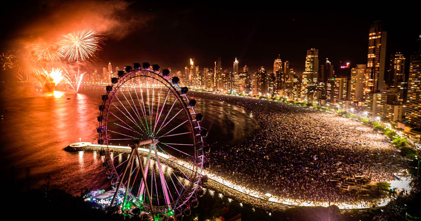 Queima de fogos em Balneário Camboriú durante o Réveillon 2025, com destaque para a roda-gigante iluminada e a orla lotada de pessoas celebrando o ano novo.