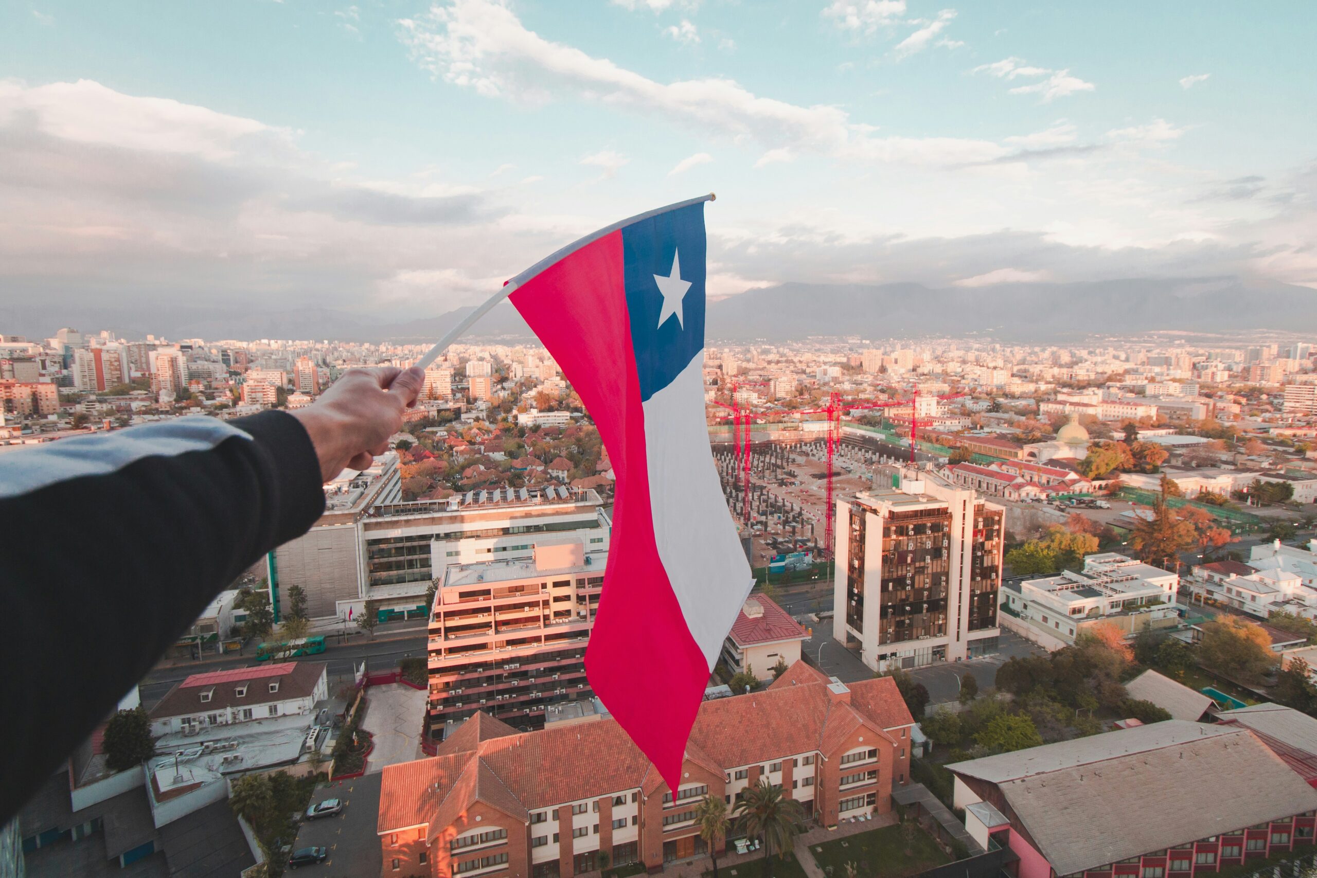 Vista aérea de Santiago, Chile, com destaque para a bandeira chilena sendo segurada em primeiro plano.