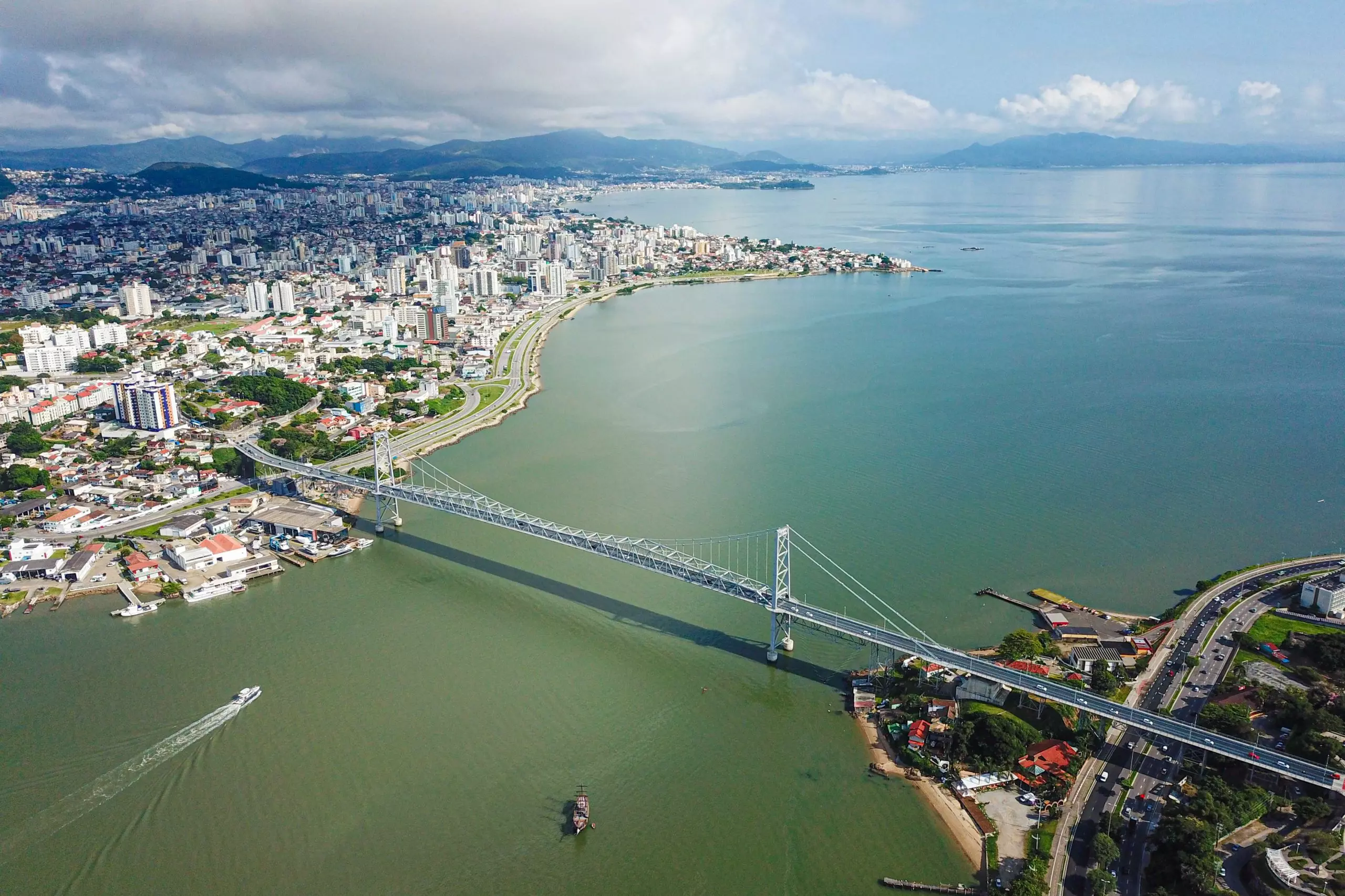 Vista aérea de Florianópolis destacando a Ponte Hercílio Luz, o mar e a cidade ao fundo.