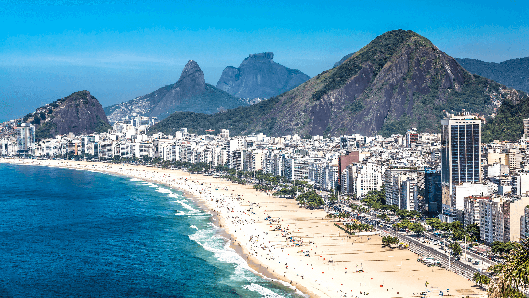 Vista panorâmica da Praia de Copacabana, Rio de Janeiro, com prédios à beira-mar e o Pão de Açúcar ao fundo, cenário perfeito para o Réveillon 2025.