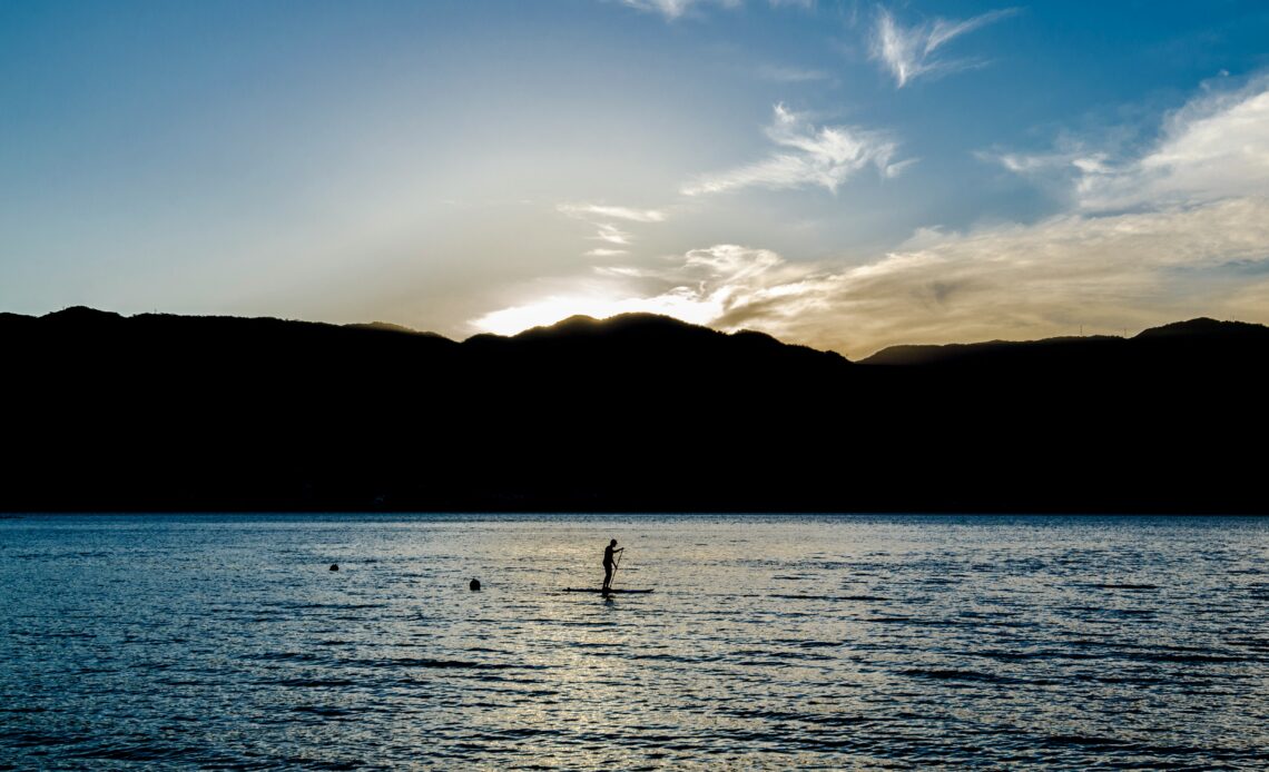 Pessoa praticando stand-up paddle ao entardecer em um lago rodeado por montanhas, simbolizando tranquilidade e aventura, ideal para viagens em 2025.