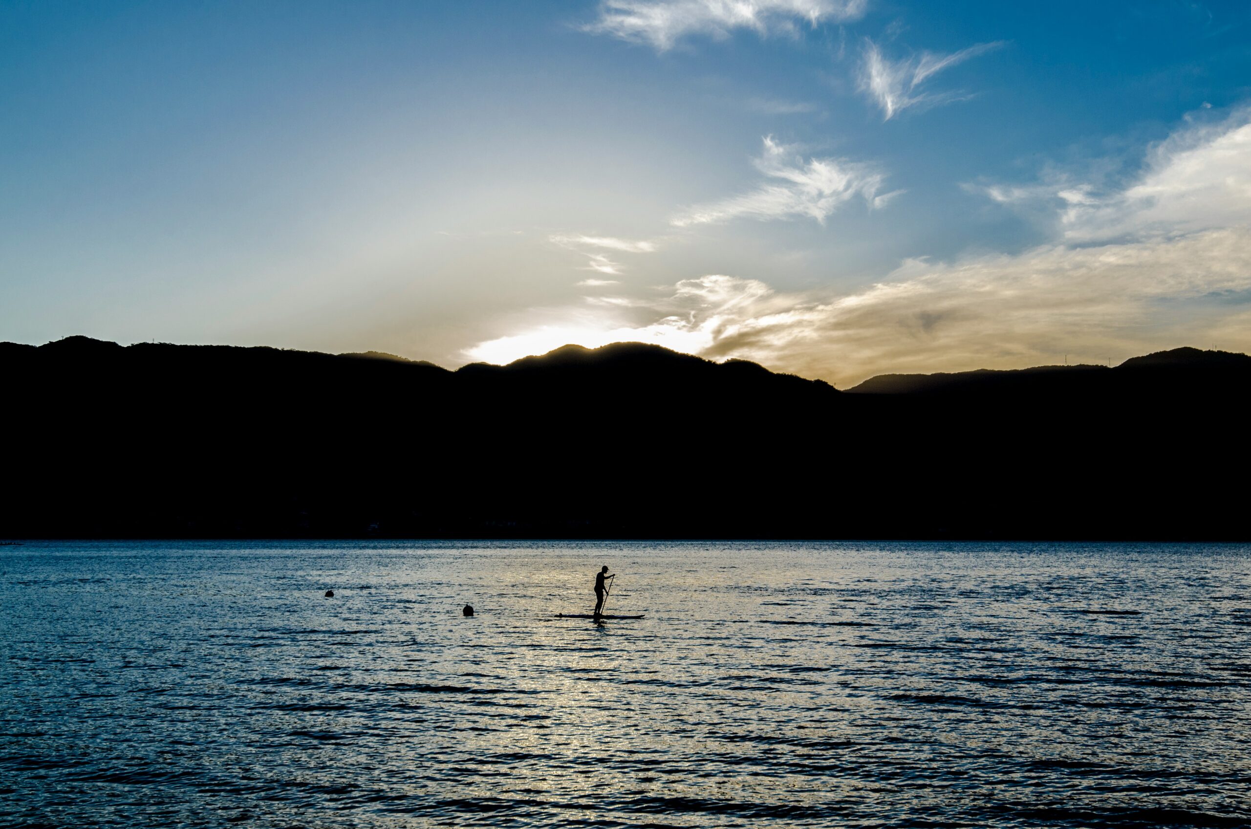 Pessoa praticando stand-up paddle ao entardecer em um lago rodeado por montanhas, simbolizando tranquilidade e aventura, ideal para viagens em 2025.