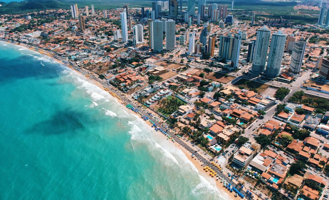 Vista aérea da Praia de Ponta Negra e sua orla urbanizada em Natal (RN), com o mar azul e prédios modernos.