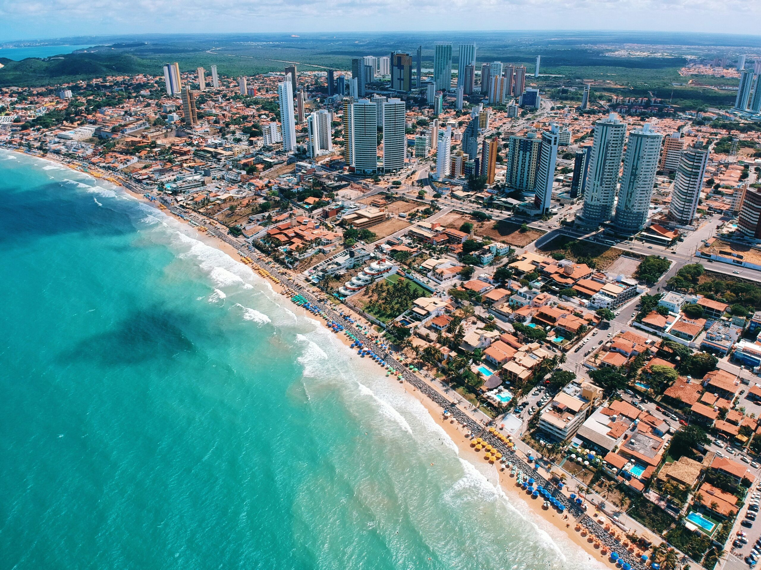 Vista aérea da Praia de Ponta Negra e sua orla urbanizada em Natal (RN), com o mar azul e prédios modernos.