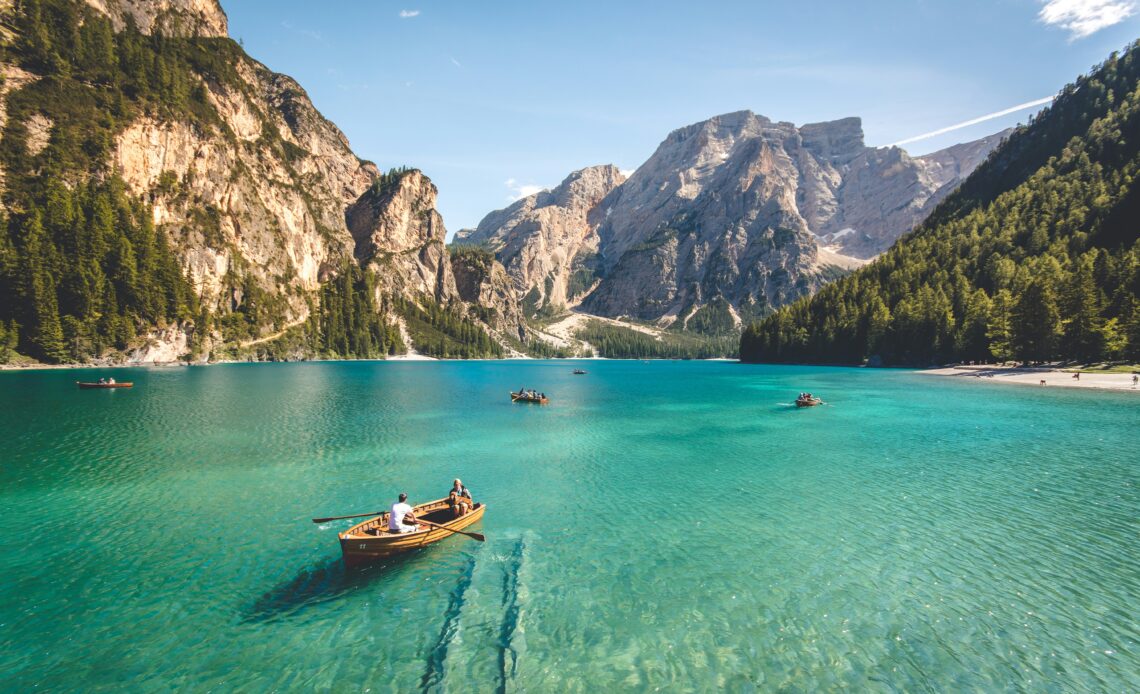 Lago de águas cristalinas cercado por montanhas rochosas e florestas, com pessoas remando em pequenos barcos de madeira. Melhores do Ano