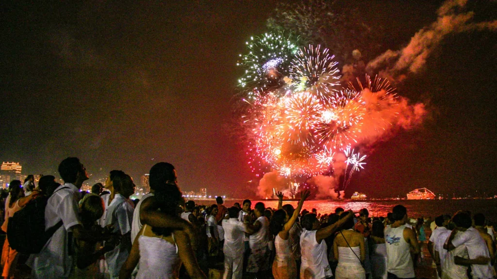 Pessoas vestidas de branco celebram o Réveillon na Praia de Copacabana, com fogos de artifício coloridos no céu.