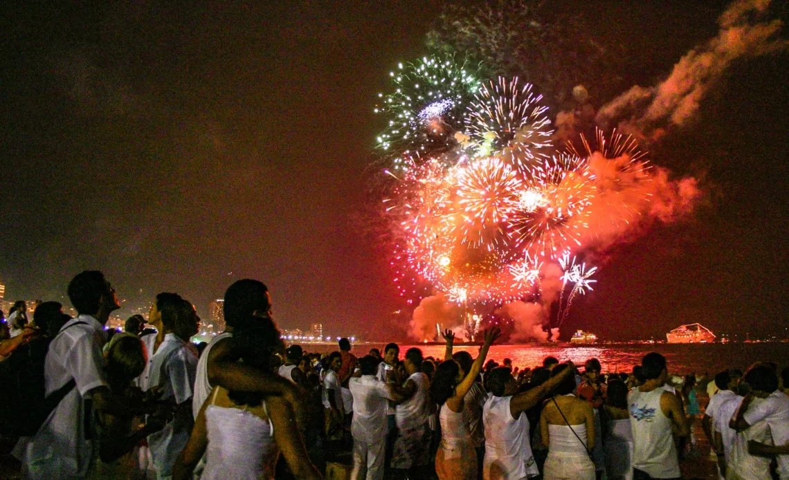 Pessoas vestidas de branco celebram o Réveillon na Praia de Copacabana, com fogos de artifício coloridos no céu.