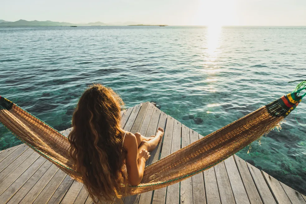 Mulher relaxando em uma rede sobre o mar durante o pôr do sol, simbolizando destinos preferidos de 2024.